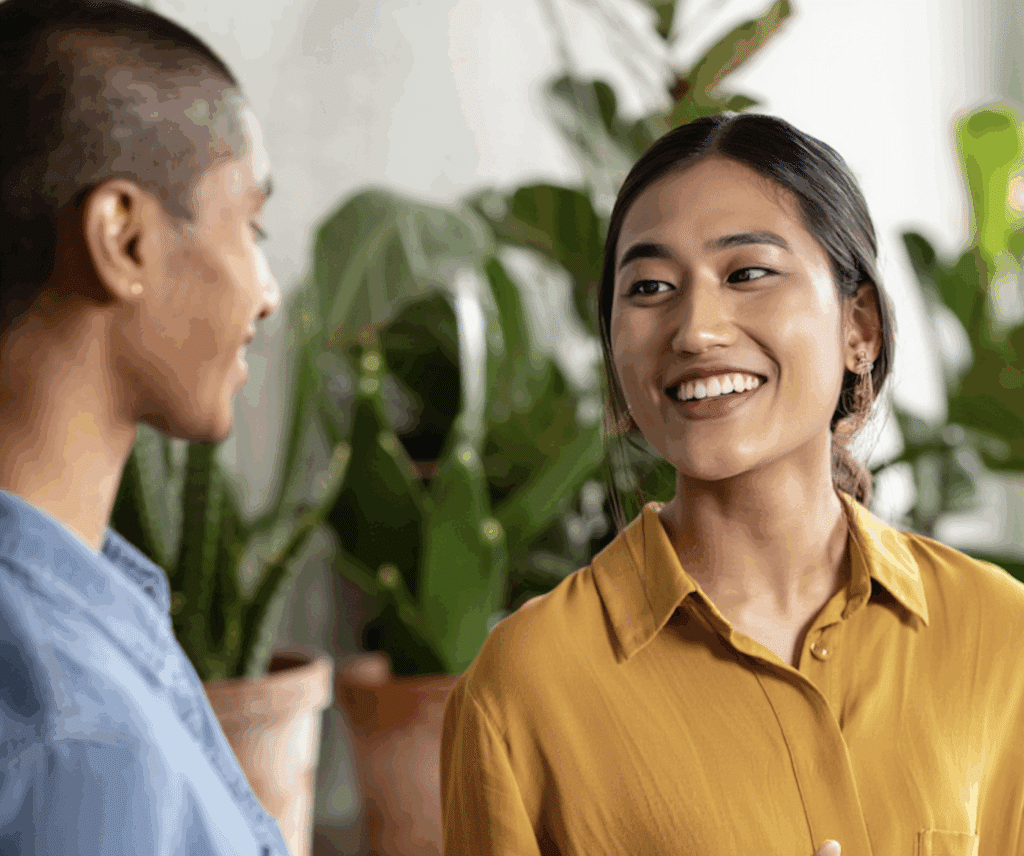 Two people are smiling and talking indoors, surrounded by green potted plants. One wears a yellow shirt and the other blue, perhaps discussing Permanent and Contract Staffing Services in a friendly, relaxed atmosphere.