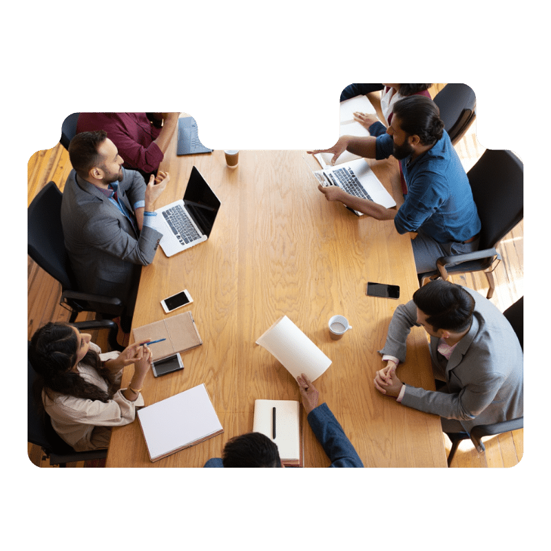A group discusses opportunities around a rectangular wooden table, laptops and papers in front of them. The meeting, viewed from above, reflects active collaboration—perfect for exploring jobs for newcomers in Toronto.