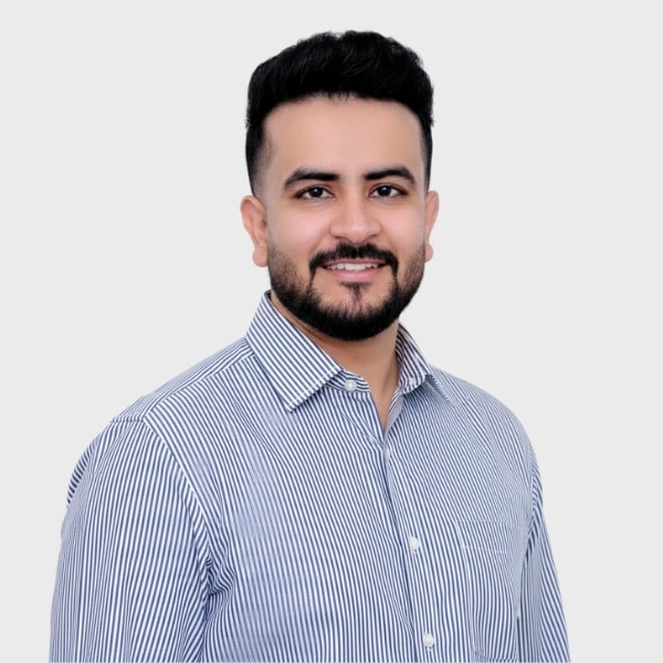 A man with short dark hair and a beard smiles at the camera. He is wearing a light blue and white striped button-up shirt, standing against a plain light gray background—perfect for an IT staffing agency in Toronto profile.