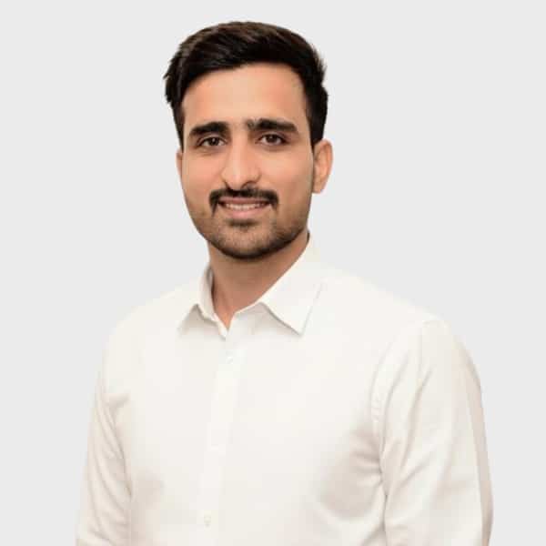 A man with short dark hair and a trimmed beard is smiling, wearing a white button-up shirt and standing against a plain light gray background, representing an IT staffing agency in Toronto.