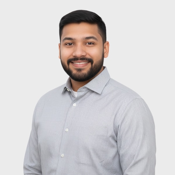 A man with short dark hair and a beard, wearing a light gray button-up shirt, smiles while standing against a plain white background, representing an IT staffing agency in Toronto.