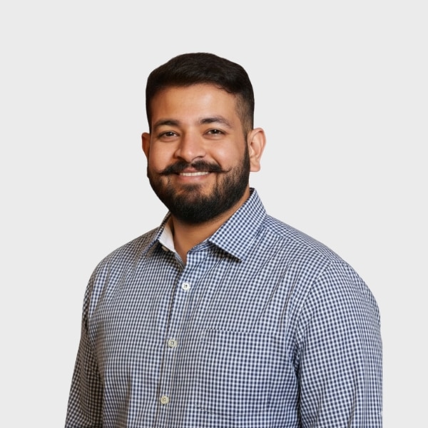 A man with short dark hair, a beard, and a handlebar mustache is smiling. He is wearing a blue and white checkered button-up shirt, standing against a plain light background—perfect for an IT staffing agency in Toronto profile photo.