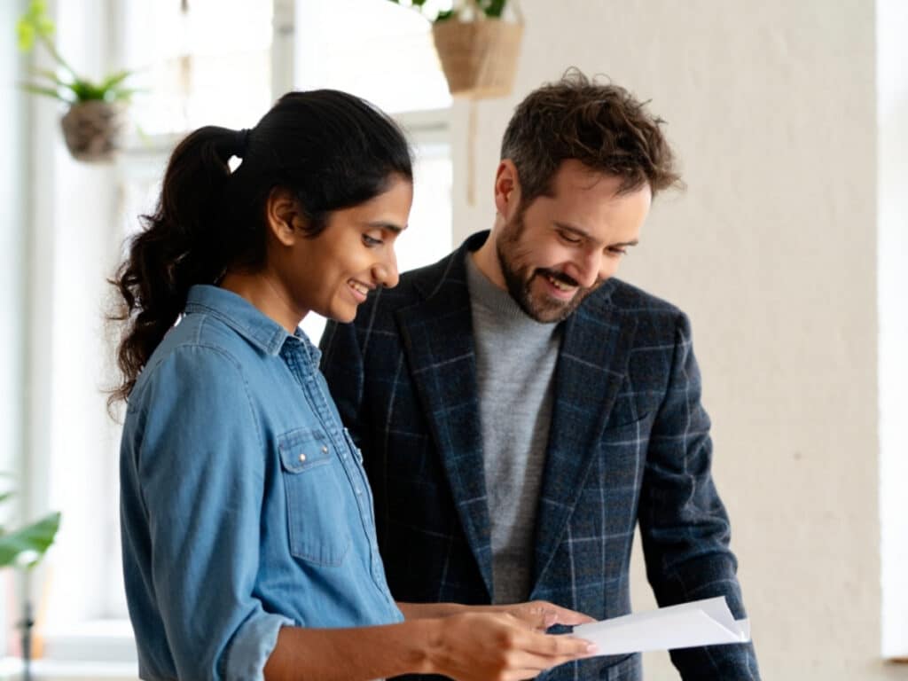 Two people standing indoors, smiling and looking at a document together—perhaps discussing IT and Finance jobs in Toronto. One wears a blue shirt, the other a plaid blazer, with bright natural light and hanging plants in the background.