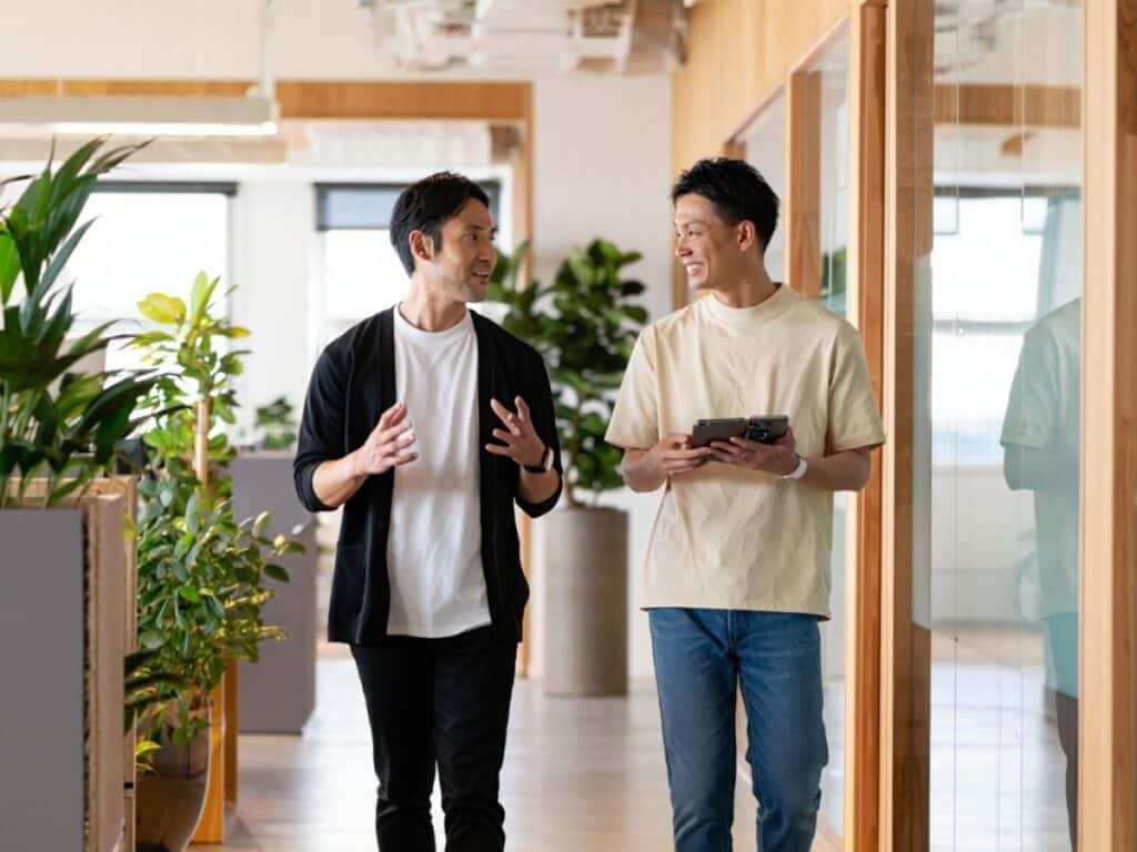 Two men discuss IT and Finance jobs in Toronto as they walk down a modern office hallway. One gestures animatedly while the other holds a phone. Green plants line the corridor, lit by sunlight streaming through large windows.