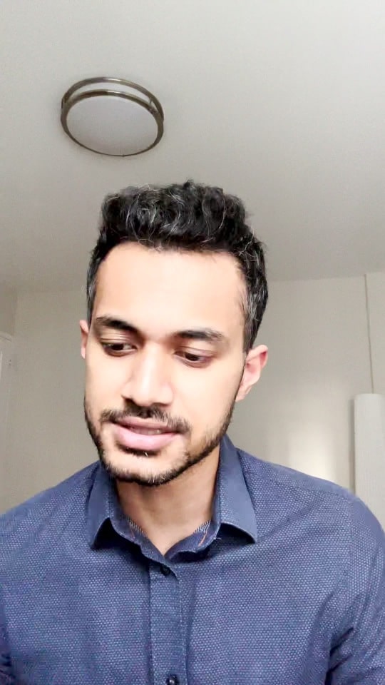 A man with short dark hair and a trimmed beard, wearing a blue button-up shirt, looks slightly down and to the side in a well-lit room—perhaps preparing for interviews for IT and Finance jobs in Toronto.