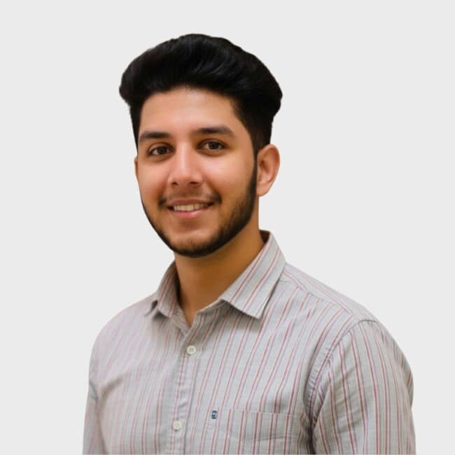 A young man with dark hair and a trimmed beard smiles at the camera. He is wearing a light-colored, striped button-up shirt, ready for his next opportunity with an IT staffing agency in Toronto, and stands against a plain, light gray background.