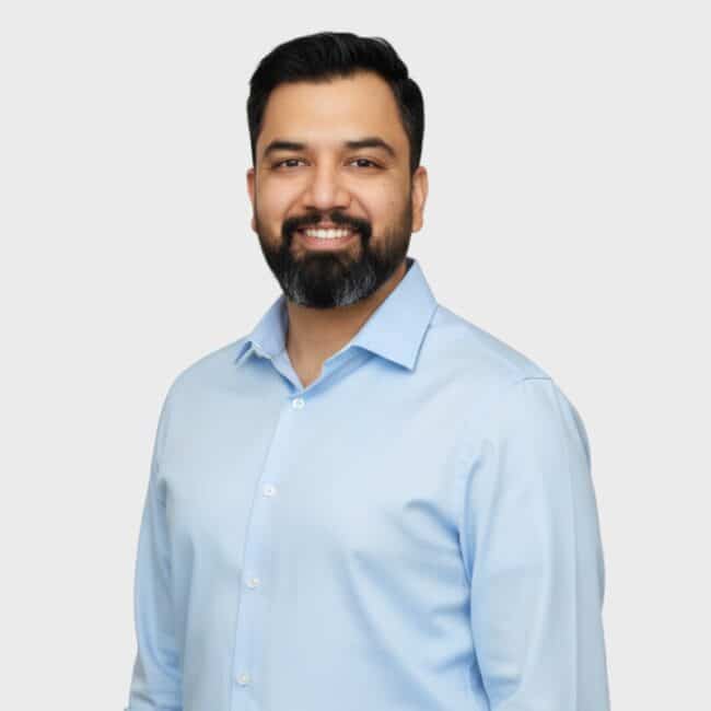 A man with dark hair and a beard smiles at the camera. He is wearing a light blue button-up shirt and stands against a plain, light gray background, representing an IT staffing agency in Toronto.