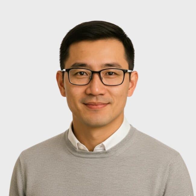 A man with short black hair and glasses, wearing a light gray sweater over a white collared shirt, smiles warmly against a plain light gray background, reflecting the professionalism of an IT staffing agency in Toronto.