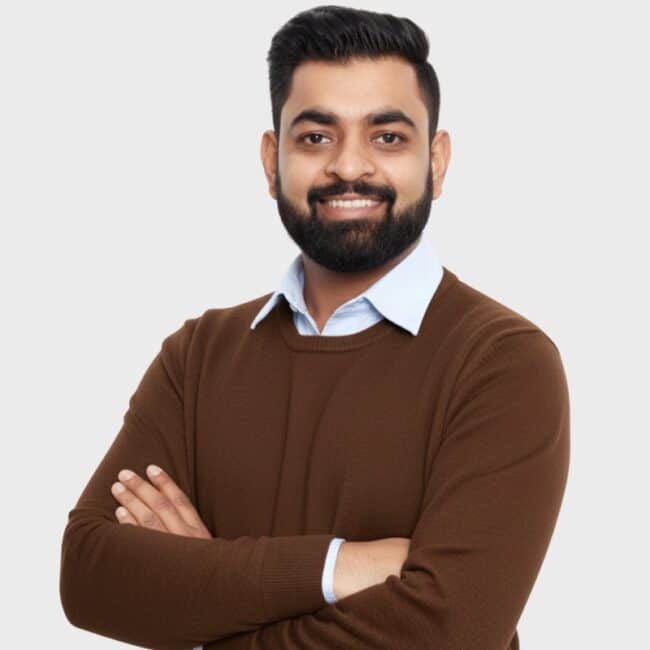 A man with dark hair and a beard smiles with his arms crossed. He wears a light blue collared shirt under a brown sweater, standing against a plain background—an approachable professional for an IT staffing agency in Toronto.