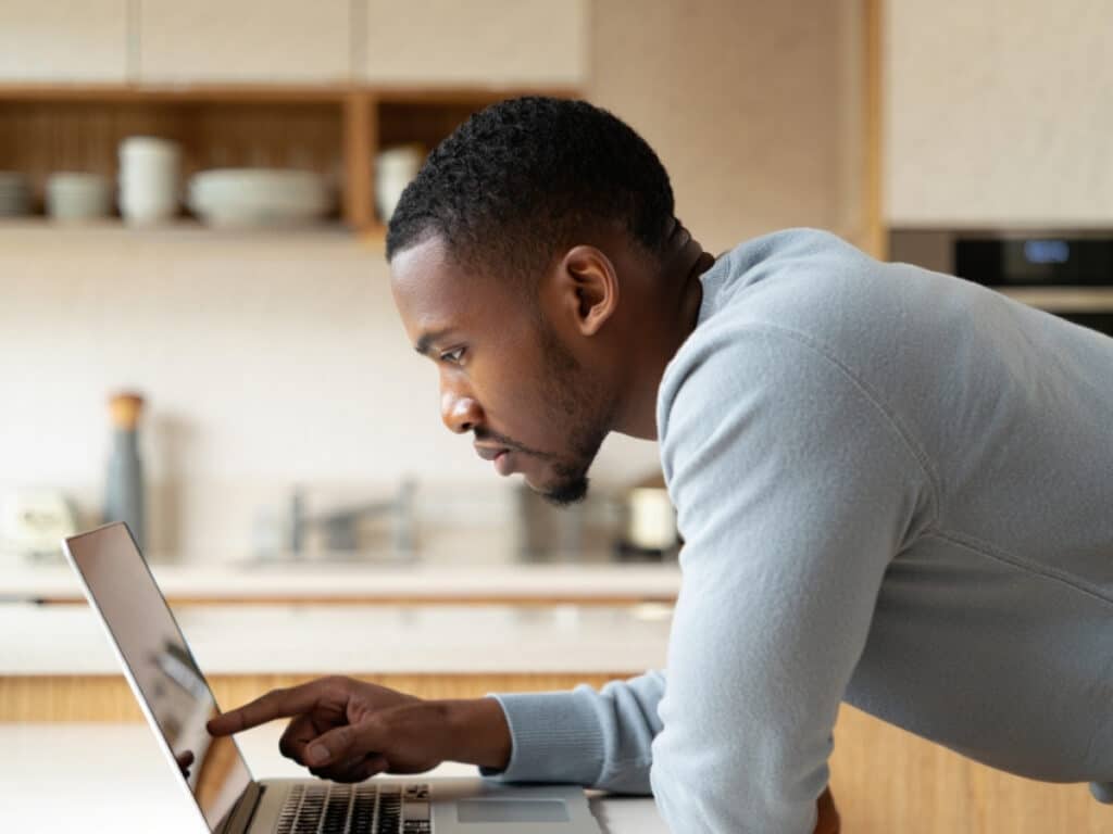 A man leans over a kitchen counter, intently looking at a laptop screen and pointing at it with one finger. Shelves with dishes and kitchen items are visible in the background.